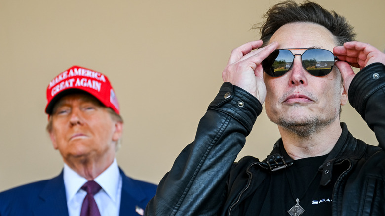 Elon Musk in sunglasses standing in front of Donald Trump in MAGA hat