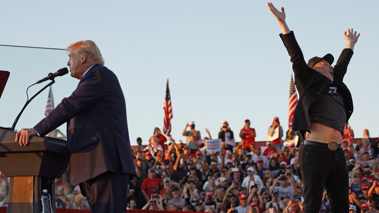 Donald Trump speaking at podium while Elon Musk jumps behind him