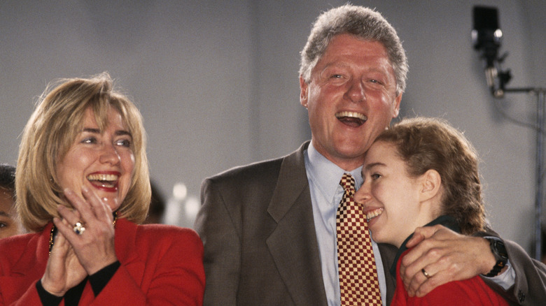 Hillary Clinton smiling and clapping while Bill Clinton laughs and hugs a smiling Chelsea Clinton