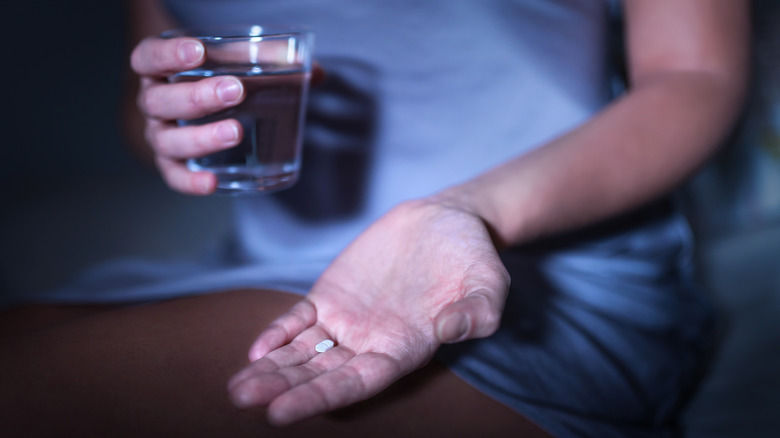 Woman sitting in a dark bedroom taking a white melatonin pill.