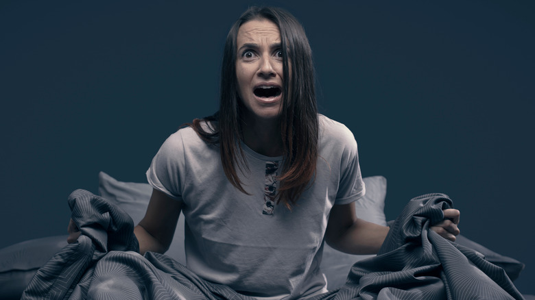 Woman wearing white t-shirt sitting in bed gripping gray sheets in frustration.