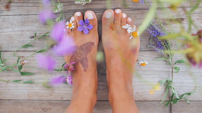 Feet on wood with white nail polish, a tattoo, and surrounding flowers