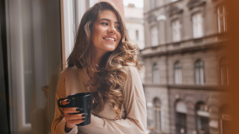 Young woman smiling while looking out the window and holding a cup of tea