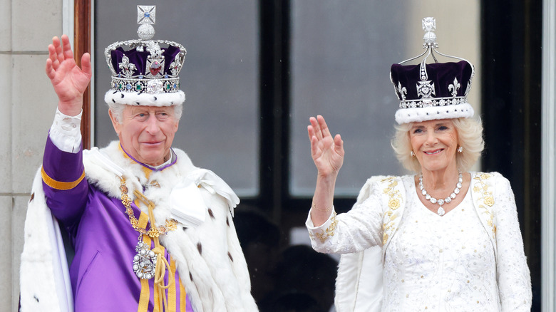 King Charles and Queen Camilla waving in crowns and robes on balcony