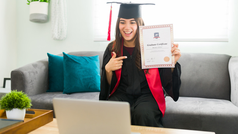 young woman in graduation outfit holding up diploma to laptop