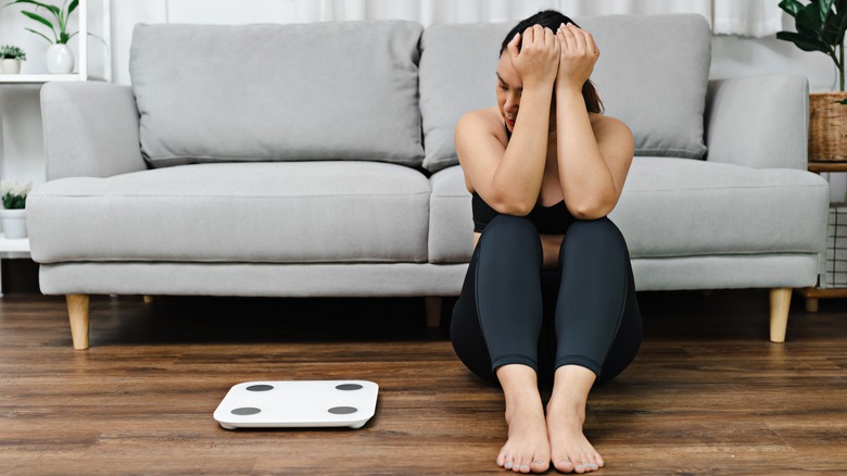 Woman frowns at a scale while sitting on the ground