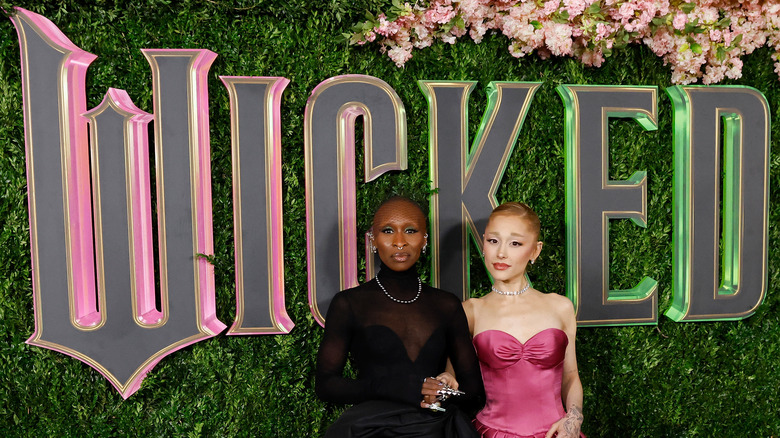 Cynthia Erivo dressed in black and Ariana Grande in pink, standing in front of a giant Wicked sign.