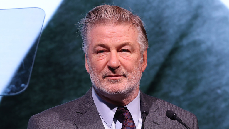 Alec baldwin stands at podium in gray suit