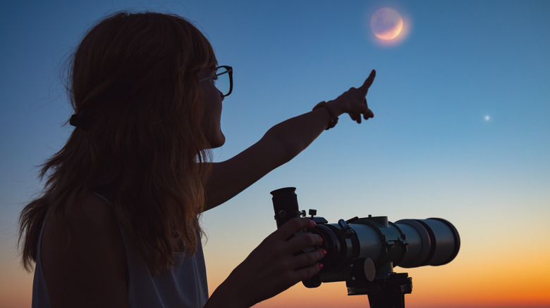 A woman pointing at the moon beside a telescope 