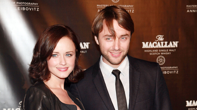 Alexis Bledel and Vincent Kartheiser smiling next to each other in front of a brown step and repeat backdrop.