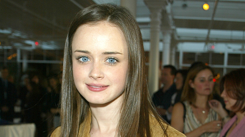 A young Alexis Bledel smiling into the camera as a crowd mingles behind her.