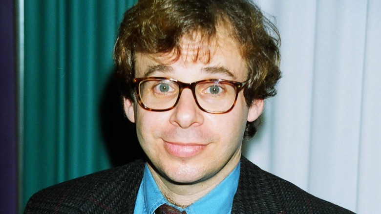 Rick Moranis wearing a blue shirt in front of green and white curtains