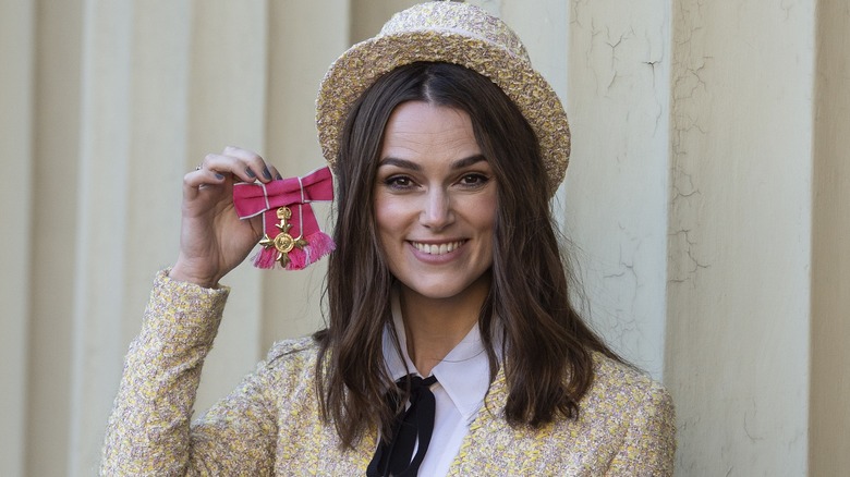 Keira Knightley in a matching bowler hat and jacket, holding up her pink OBE