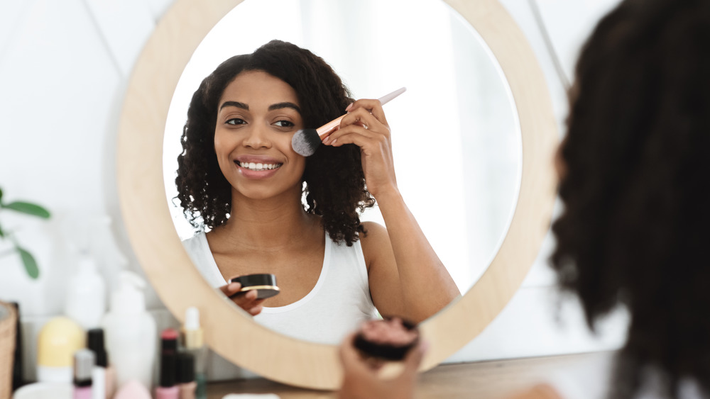 A woman is smiling and facing a round mirror while applying blush to her cheek.