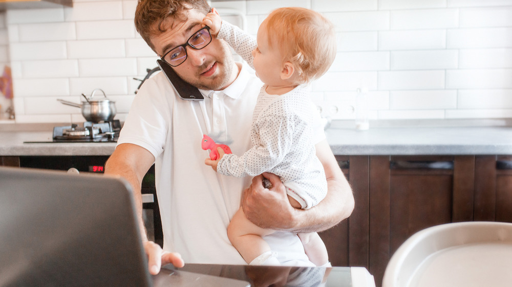 A man typing on a laptop while a baby reaches for his face