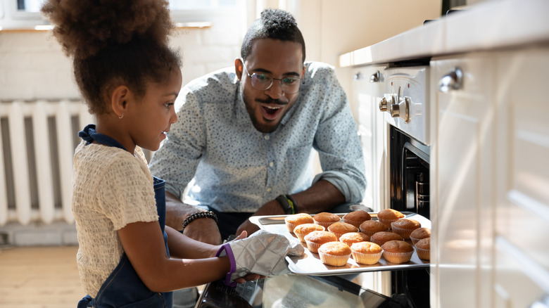 Girl taking muffins out of the oven while her father watches