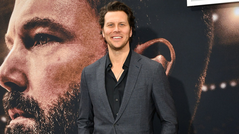 Hayes MacArthur standing and smiling in front of a giant Ben Affleck poster