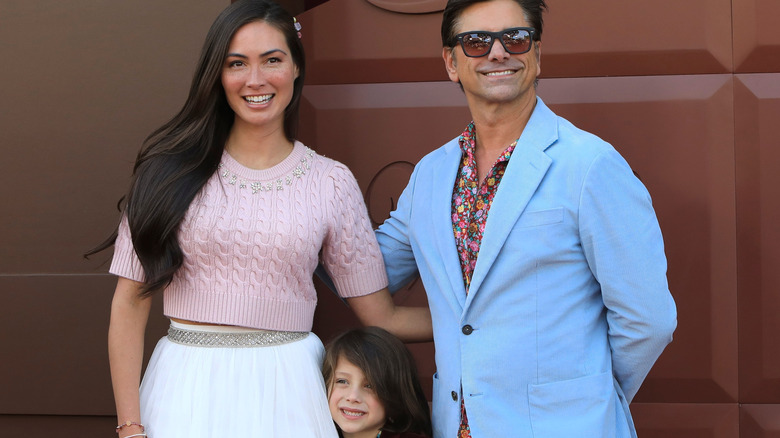 John Stamos stands smiling next to his wife with their son in between them against brown background