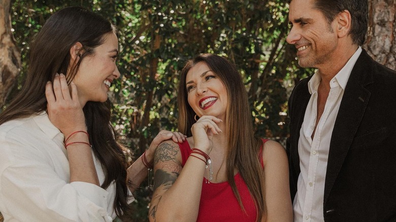 Caitlin McHugh and John Stamos look at a woman in the middle of them laughing and modeling red bracelets outside