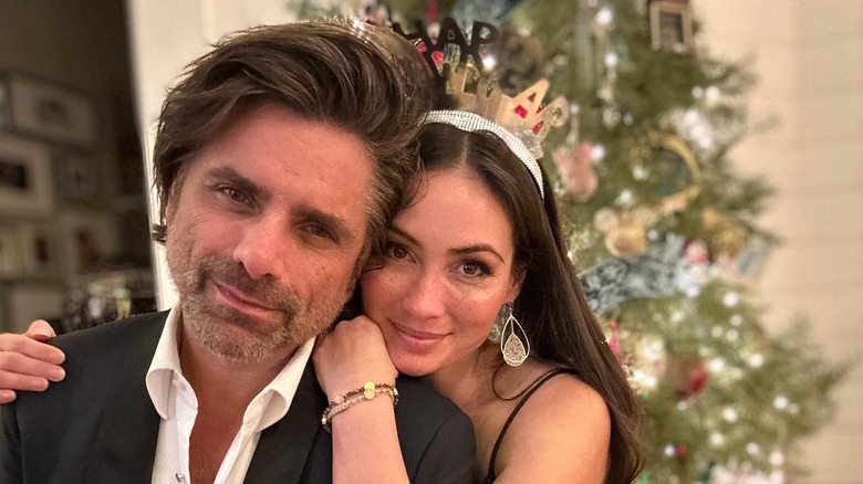 Caitlin McHugh has arms around John Stamos smiling in front of a christmas tree for NYE