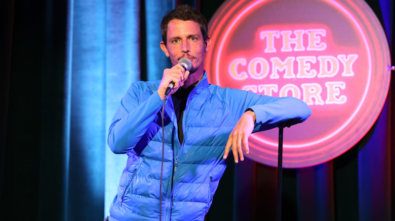 Tony Hinchcliffe leaning on a microphone stand in a blue jacket