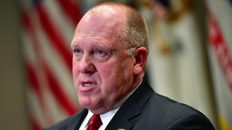 Tom Homan speaking in front of a row of American flags