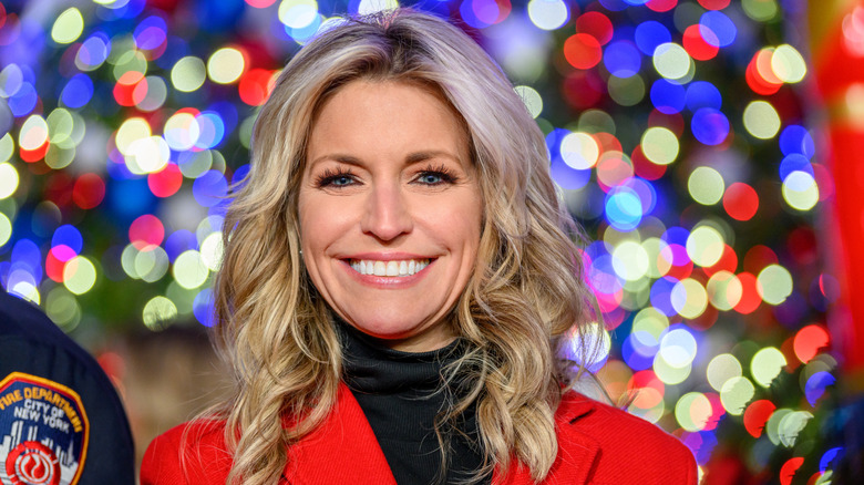 Ainsley Earhardt with in a black highneck top and a red jacket smiling as she poses in front of bright lights