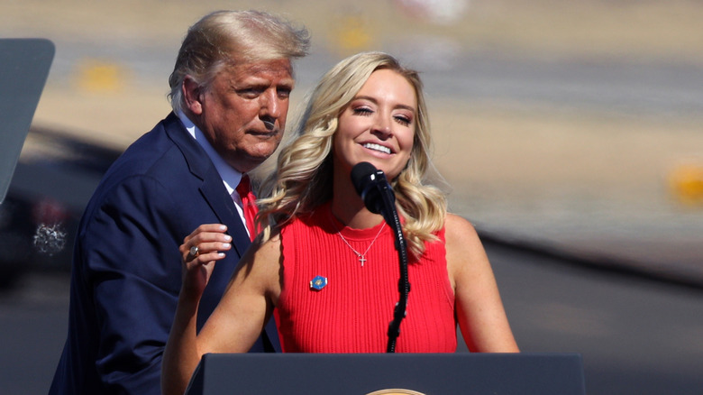 Kayleigh McEnany stands behind podium, Donald Trump stands behind her