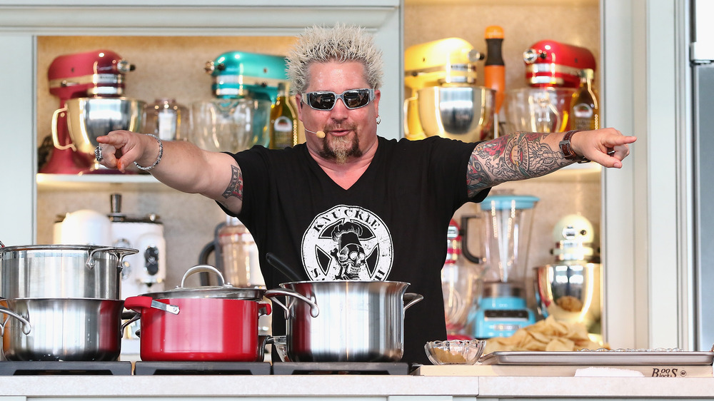 Guy Fieri wearing sunglasses and pointing both hands out with pots sitting in front of him on a stove.