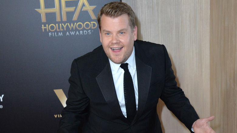 James Corden smiles in a suit and tie with his hands out.