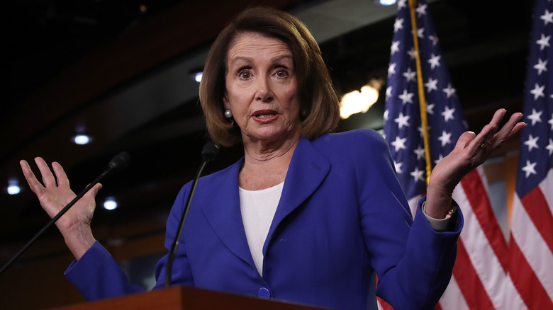 Nancy Pelosi holds her hands up at a podium with surprised expression