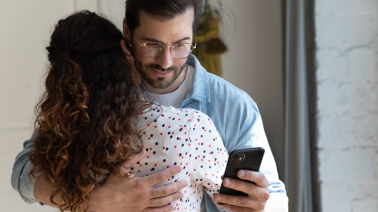 Man secretly looking at phone while hugging woman