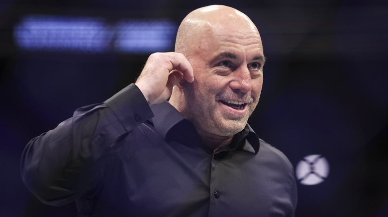 Joe Rogan in black dress shirt, hand over hear, smiling devilishly.