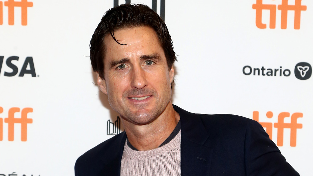 Luke Wilson wears a black blazer over a beige sweater.