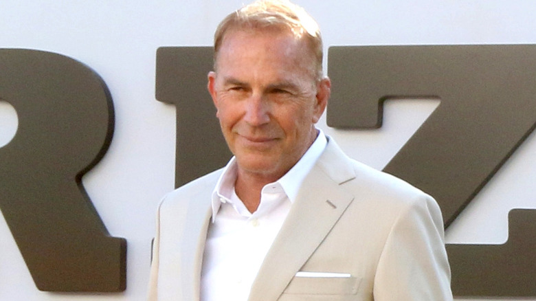 Kevin Costner on red carpet wearing white shirt and beige jacket, small smile, looking away from camera.